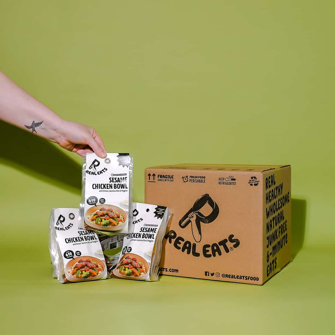 A RealEats meal kit box next to three of RealEats meals. a hand is grabbing one of the meal pouches.