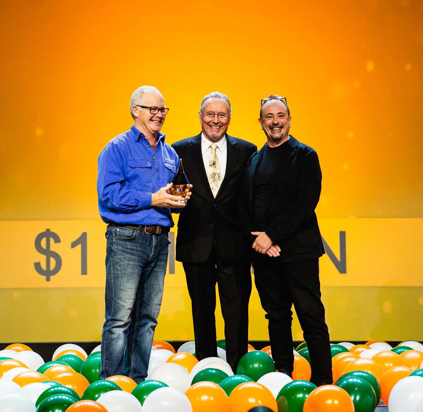 three men stand on a stage holding a prize surrounded by balloons
