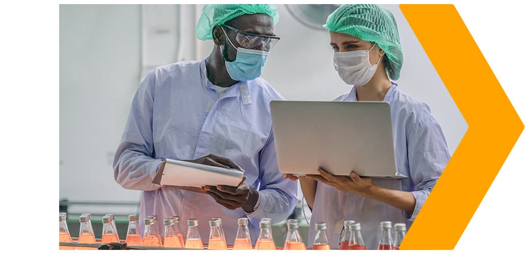 A Black man and white woman with masks and hairnets on take notes in a beverage bottling facility, and a yellow arrow points right